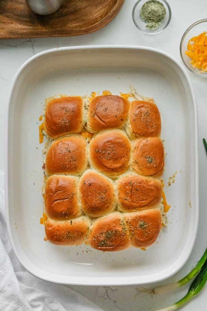 A white baking dish filled with twelve golden-brown crack chicken sliders topped with butter and herbs.