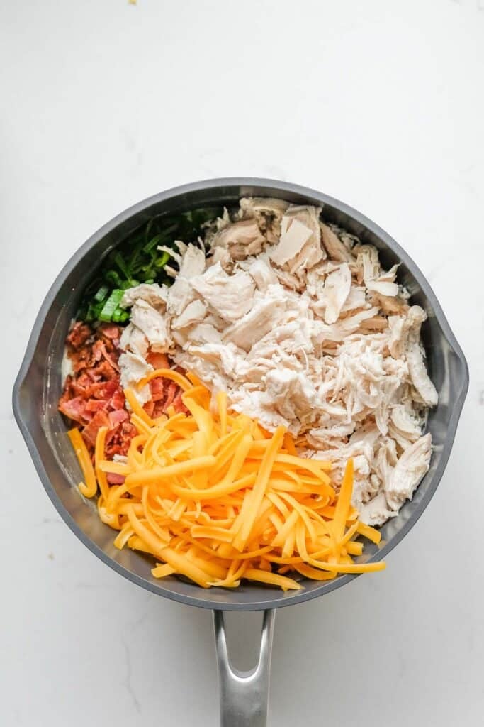 The saucepan of creamy sauce with shredded cheese, shredded chicken, bacon bits and green onions added to mixture.