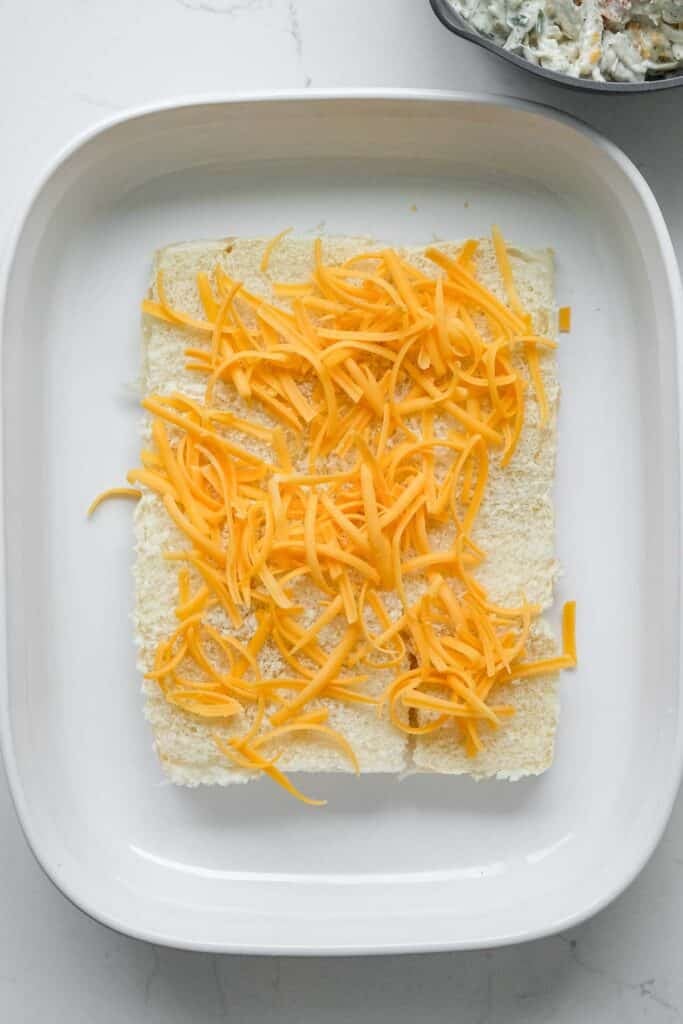 Shredded cheddar cheese is spread over bottoms of the hawaiian rolls in a white baking dish.