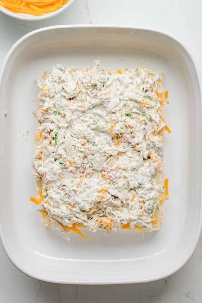 A rectangular layer of creamy chicken mixture with shredded cheese is spread over sliced bottoms in a white baking dish.