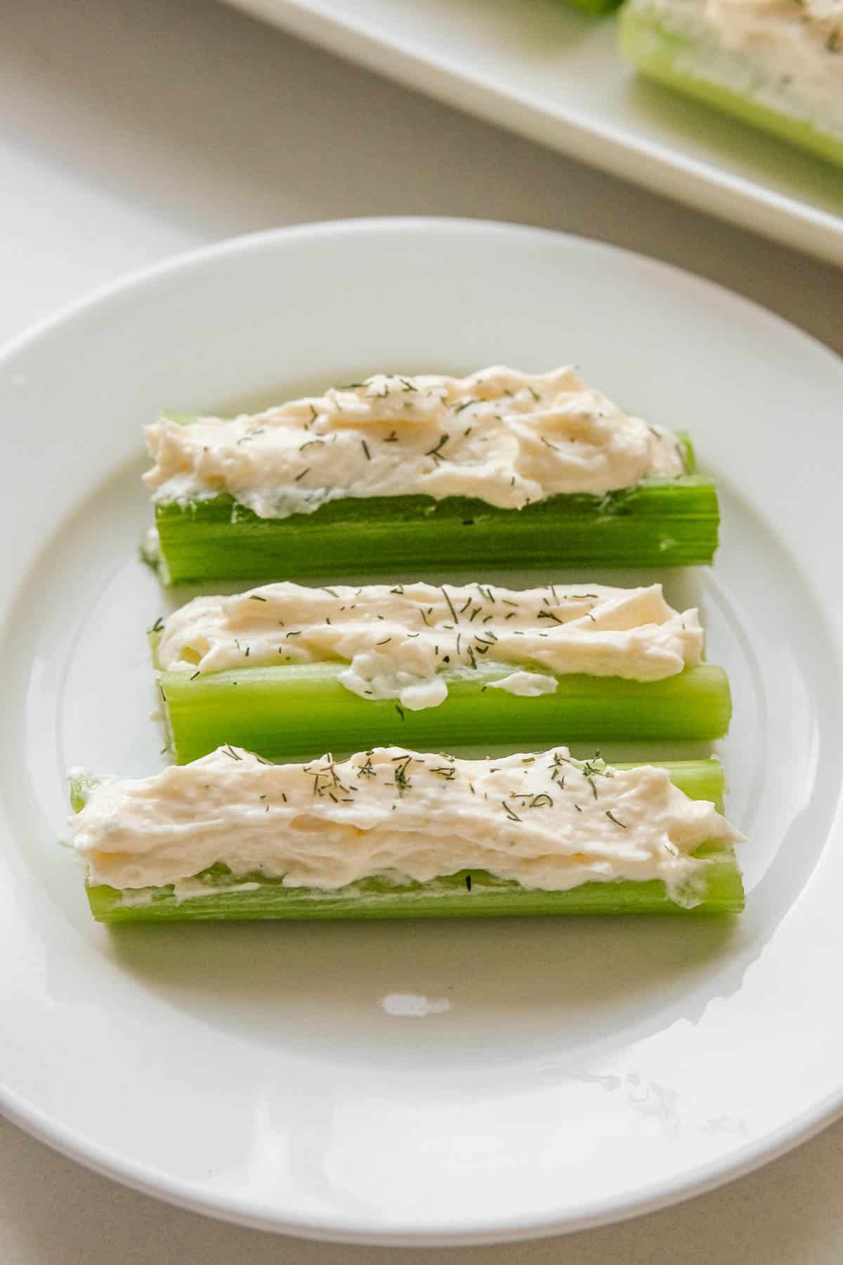 Three stuffed celery sticks filled with a cream cheese spread and sprinkled with herbs, served on a white plate.
