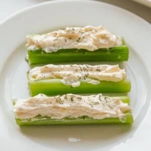Three stuffed celery sticks filled with a cream cheese spread and sprinkled with herbs, served on a white plate.