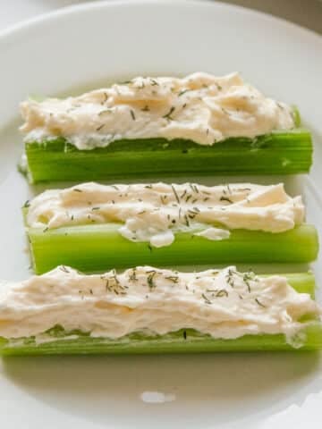 Three stuffed celery sticks filled with a cream cheese spread and sprinkled with herbs, served on a white plate.