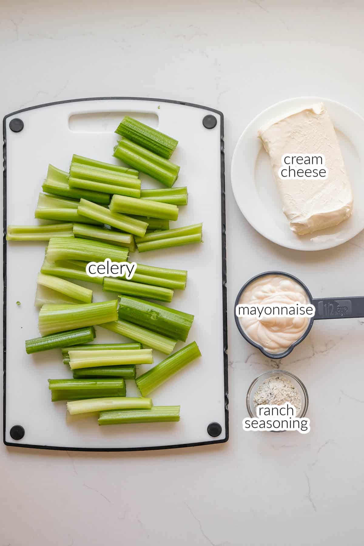 A cutting board of ingredients for stuffed celery including chopped celery sticks, a plate of cream cheese, mayonnaise, and ranch seasoning on a white countertop.