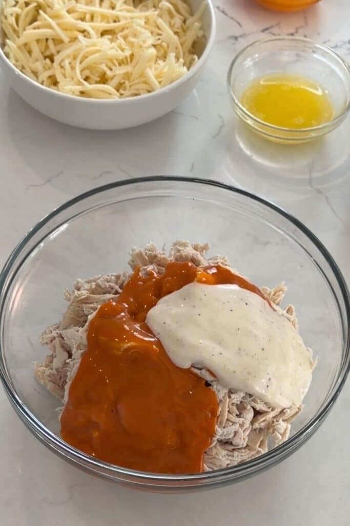 A glass bowl with shredded chicken topped with buffalo sauce and ranch dressing. Nearby are bowls of shredded cheese and melted butter on a white countertop.