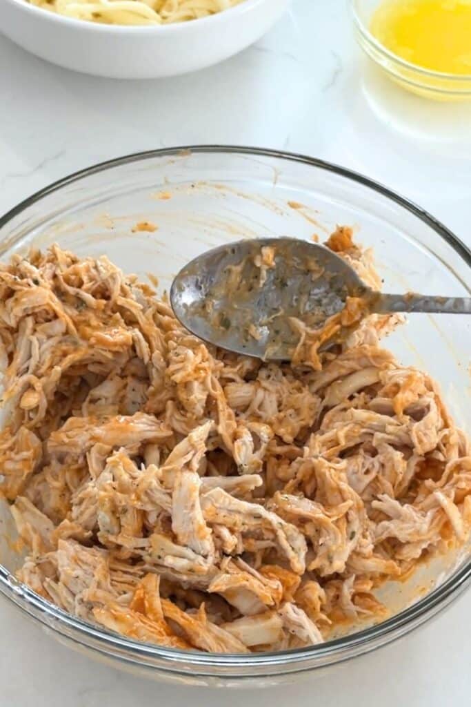 A glass bowl filled with shredded chicken mixed with buffalo sauce, with a metal spoon resting on top. A bowl of melted butter and another bowl are in the background.
