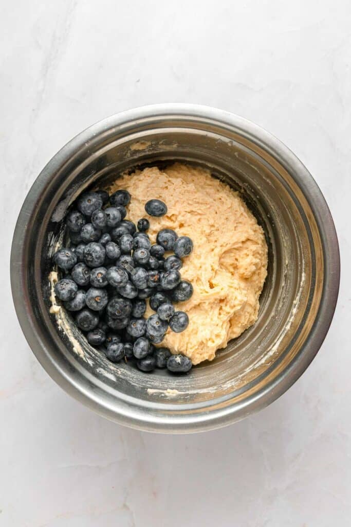 A stainless steel mixing bowl containing muffin batter with fresh blueberries on top, ready to be mixed in.