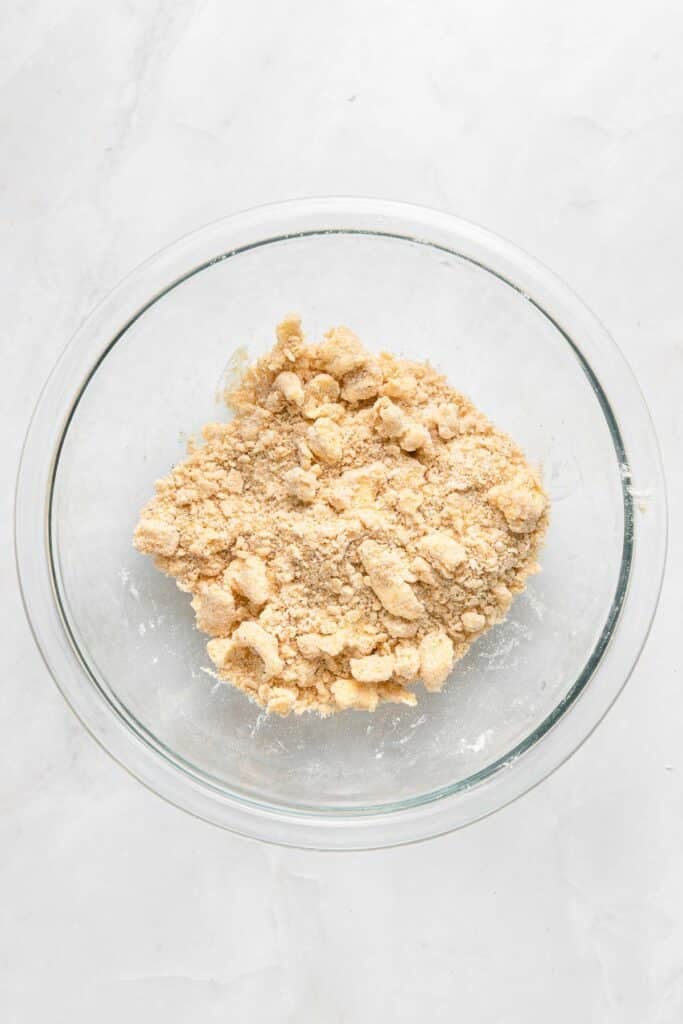 A glass bowl containing a coarse, crumbly dough mixture sits on a white surface.