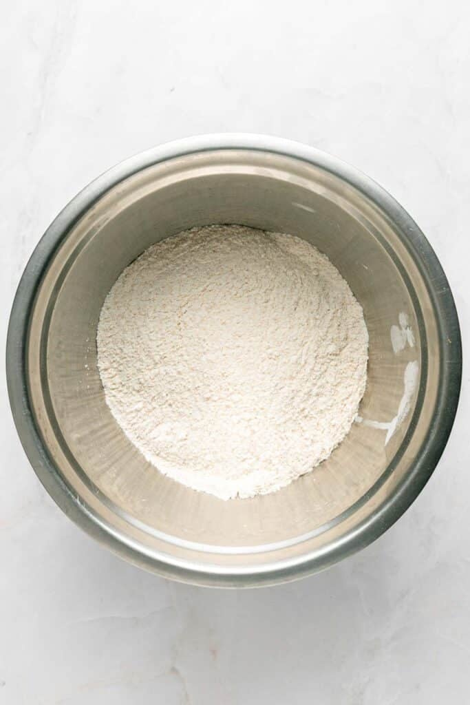 A stainless steel bowl containing a mound of white flour sits on a light-colored surface.