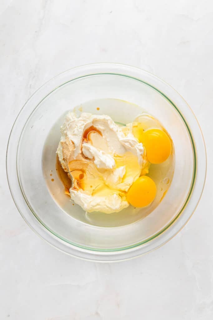 A glass bowl containing two raw eggs, greek yogurt, sugar, and vanilla extract on a white surface.