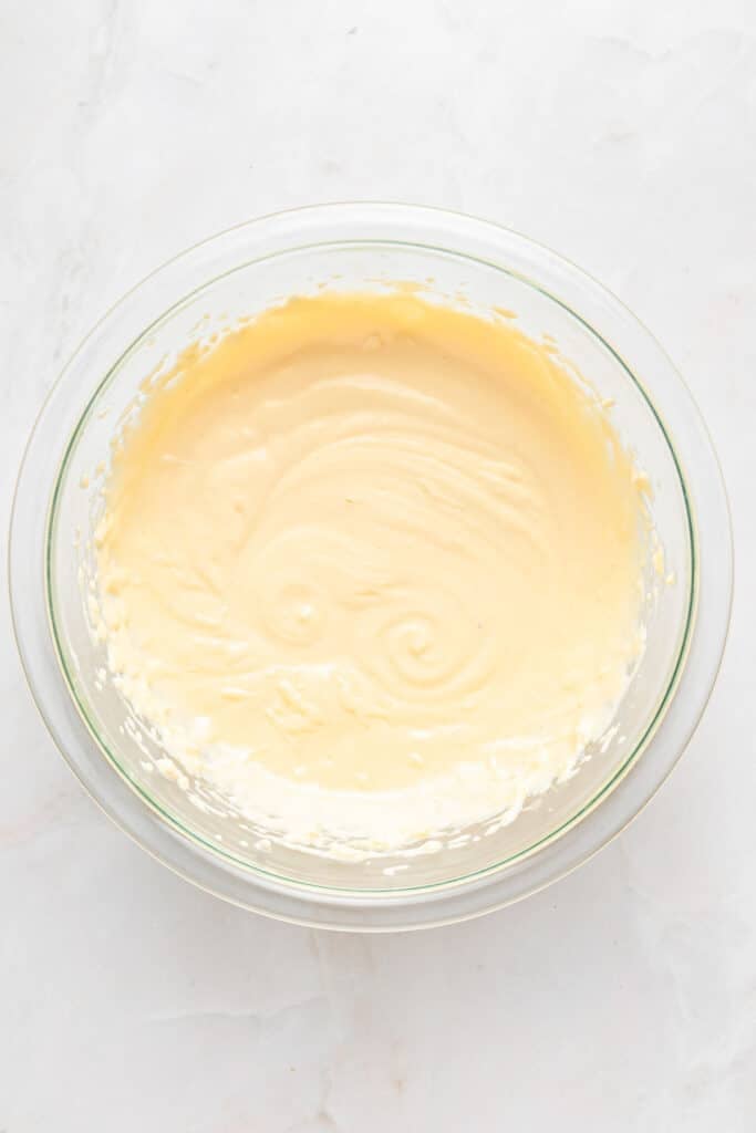 A glass bowl filled with smooth, pale yellow cake batter on a white marble surface.