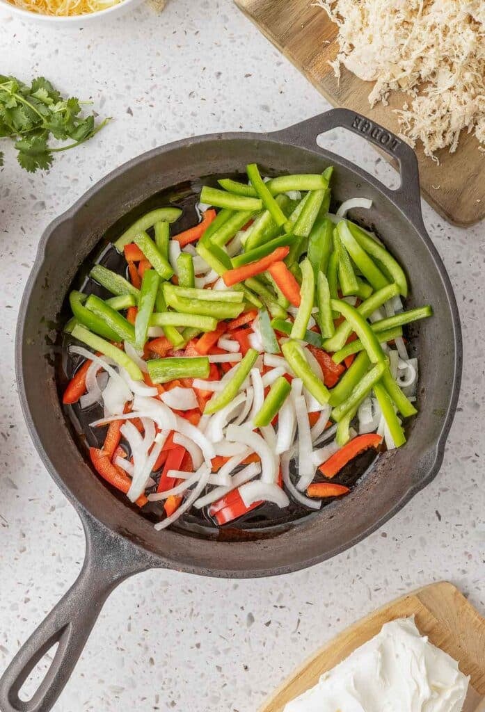 Sliced green bell peppers, red bell peppers, and onions cooking in a cast iron skillet on a light-colored countertop.