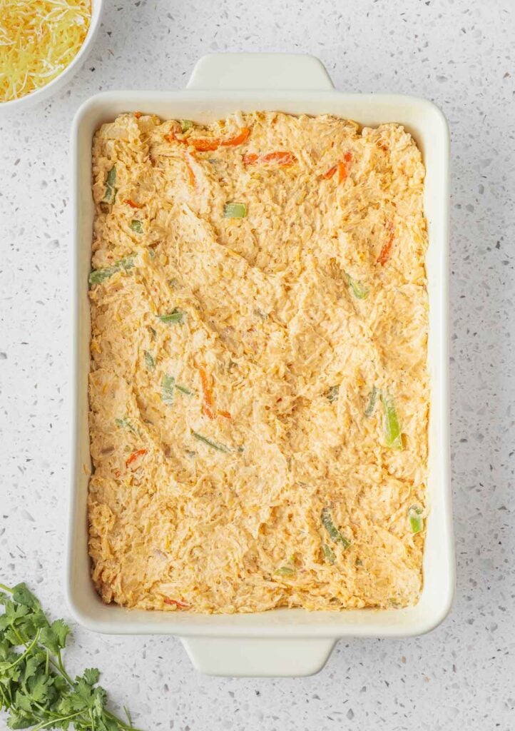 A rectangular casserole dish filled with an unbaked creamy mixture of shredded chicken, cheese, and chopped vegetables on a white countertop.