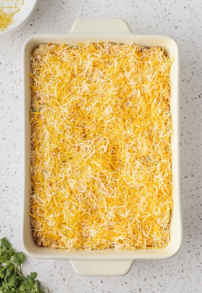 A rectangular casserole dish filled with an uncooked Chicken Fajita Casserole topped with a generous layer of shredded yellow and white cheese.