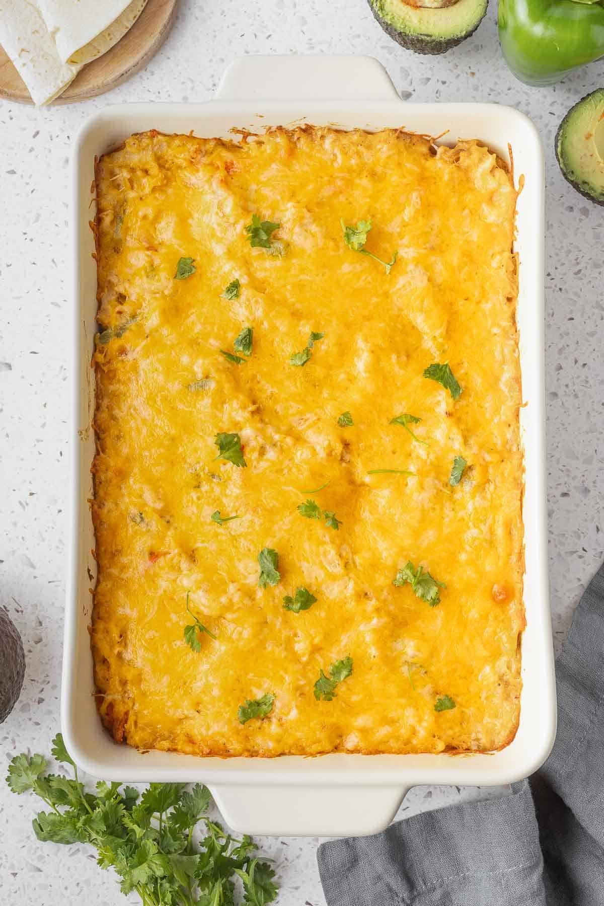 A rectangular casserole dish filled with a Chicken Fajita Casserole, garnished with small sprigs of cilantro, sits on a light countertop with avocados and cilantro nearby.