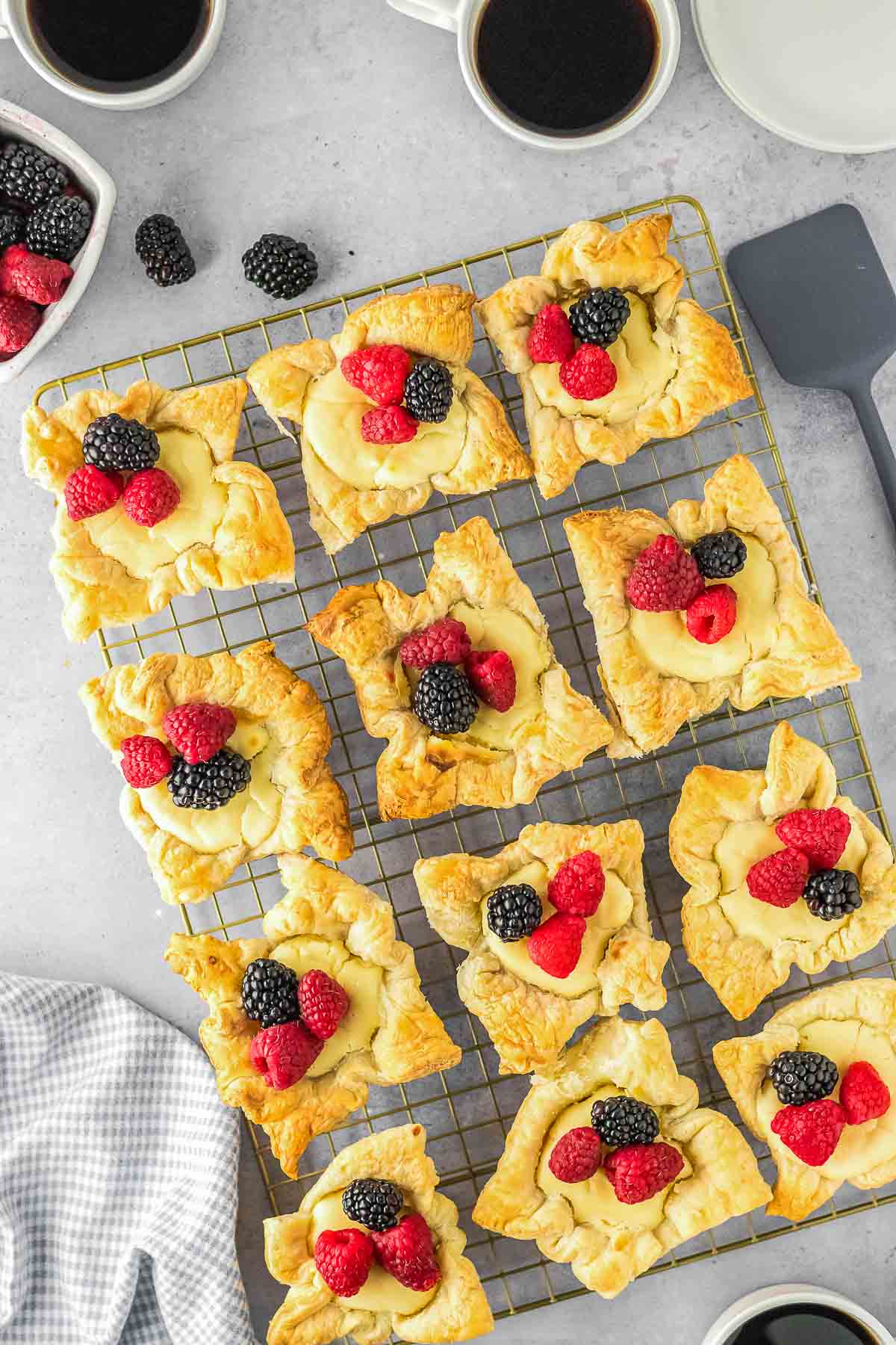 Twelve cream cheese puff pastries topped with custard and fresh raspberries and blackberries on a cooling rack, with cups of black coffee nearby.