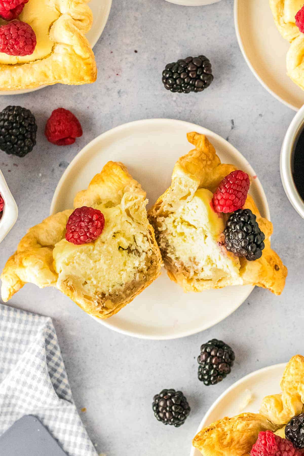 A cream cheese puff pastry cut in half on a plate, topped with a raspberry and blackberry, with more berries and pastries nearby.