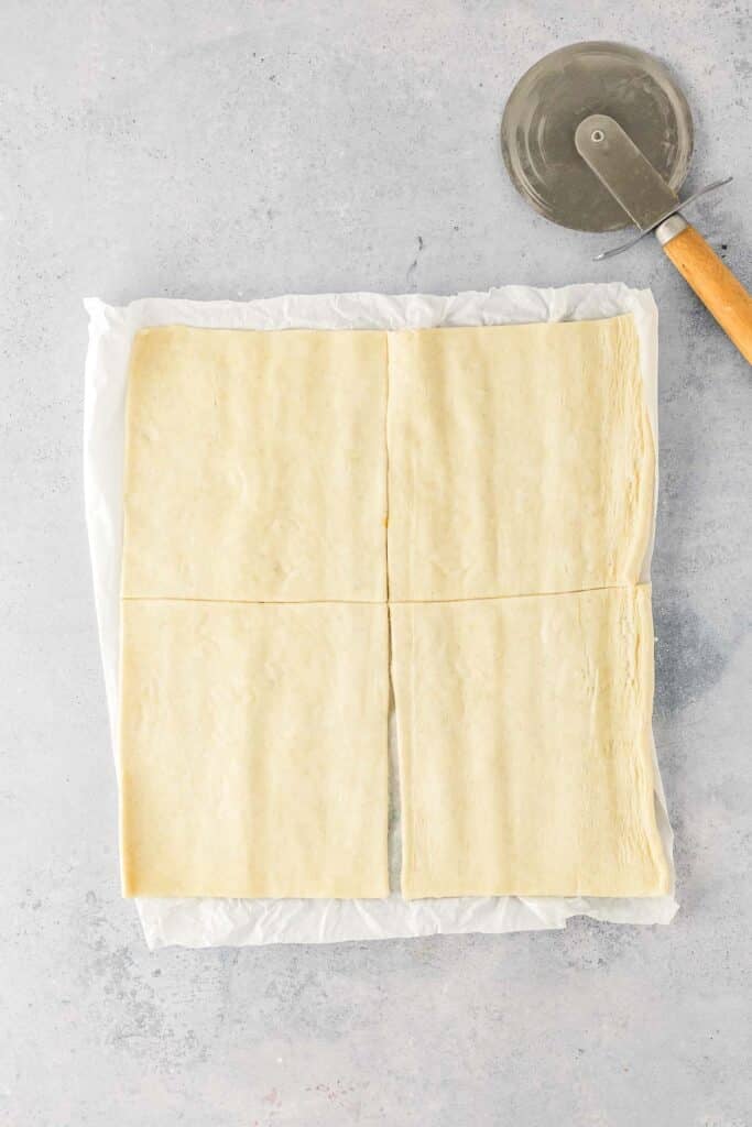 A sheet of uncooked pastry dough cut into four rectangles on parchment paper, with a pizza cutter lying next to it on a light surface.