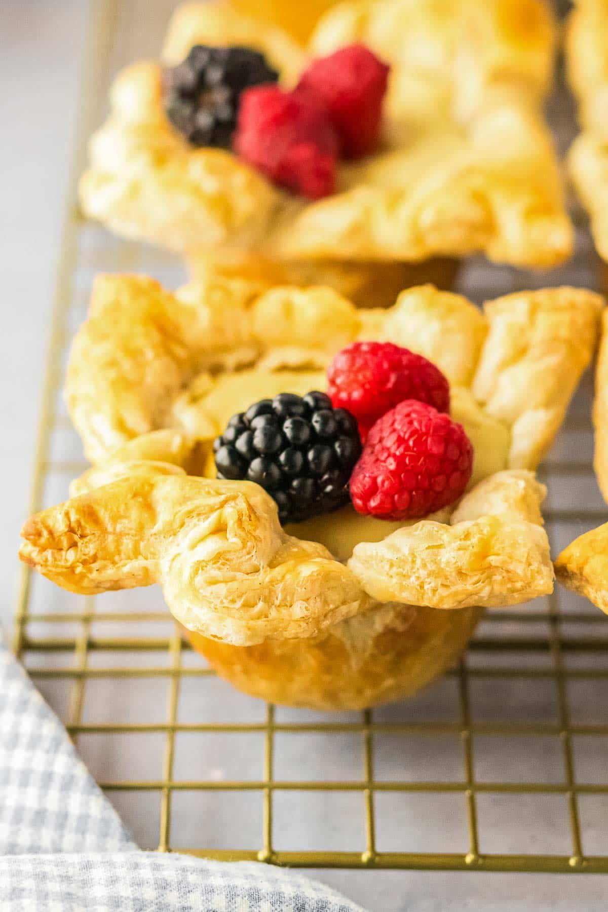 A cream cheese puff pastry with a flaky crust topped with fresh raspberries and a blackberry, sits on a cooling rack.