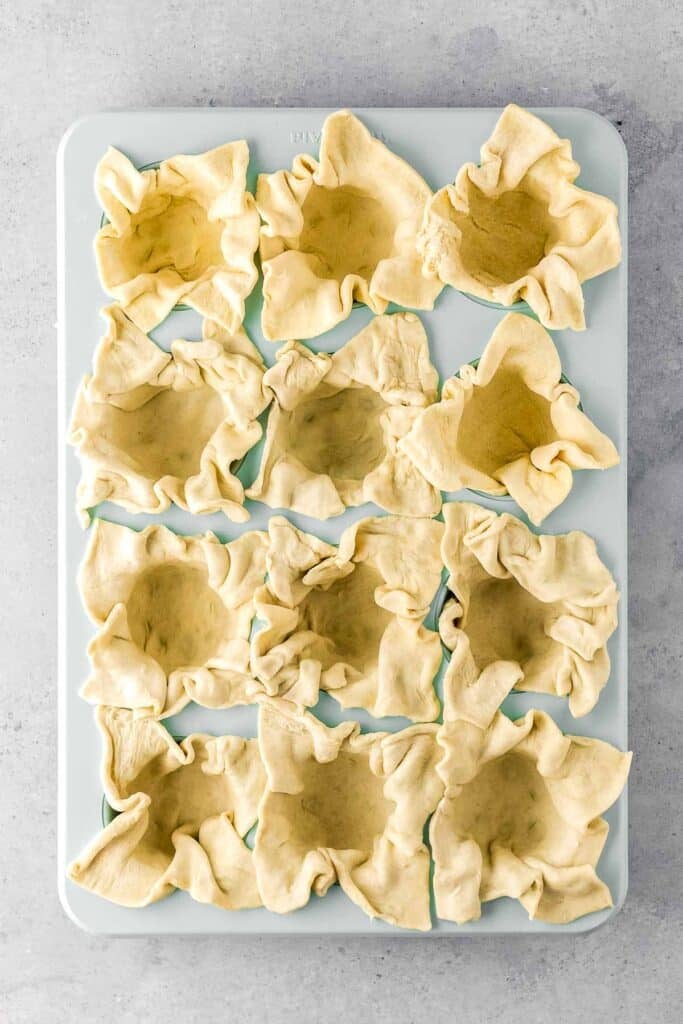 A muffin tin filled with unbaked, crimped pastry dough cups ready for filling.