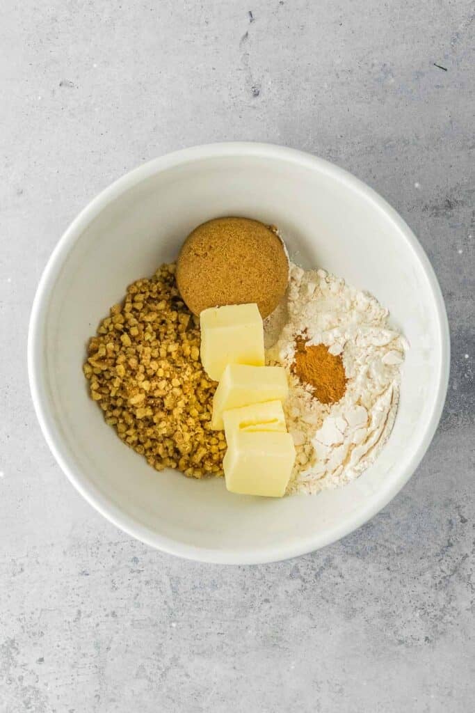 A white bowl containing chopped nuts, brown sugar, flour, cinnamon, and three chunks of butter on a light gray surface.