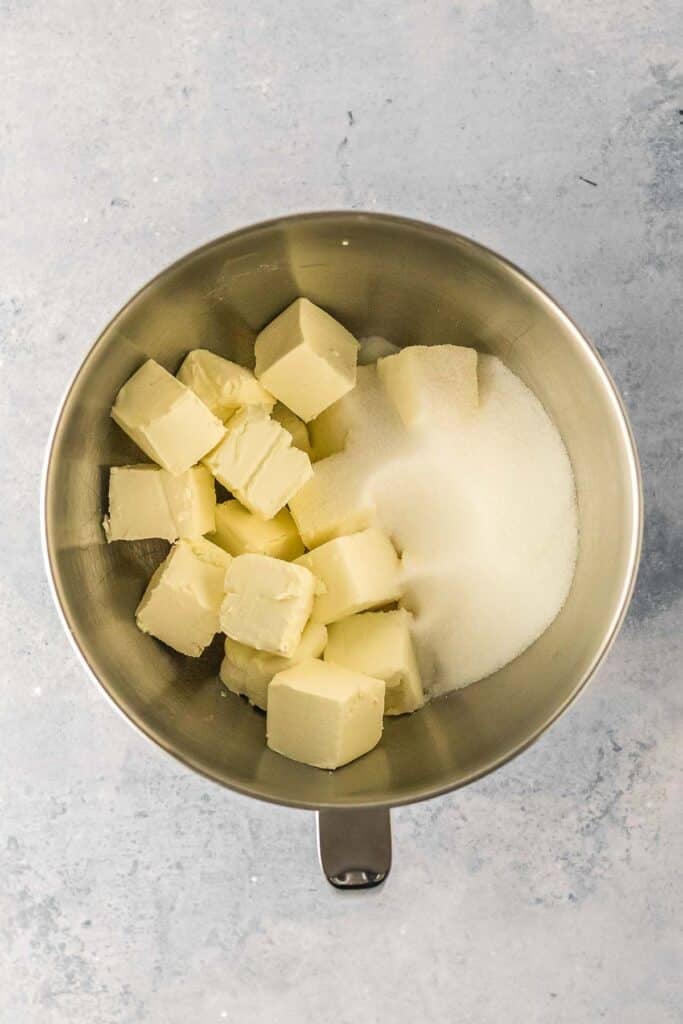 A metal mixing bowl containing cream cheese and granulated sugar on a light gray surface.