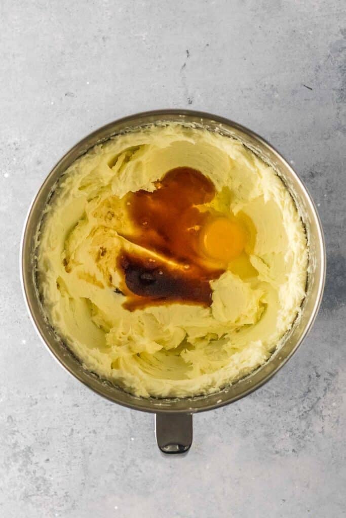 A mixing bowl containing creamed butter and sugar with an egg and vanilla extract added on top, ready to be mixed.