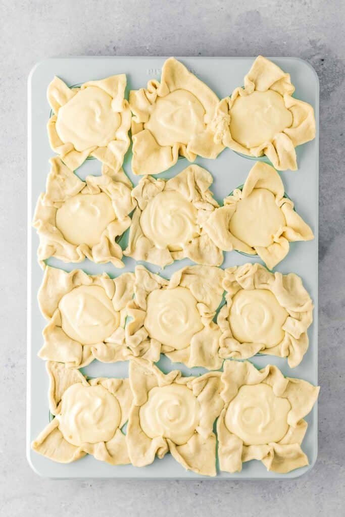A baking tray with twelve puff pastry cups filled with creamy batter, arranged in three rows on a light-colored surface.
