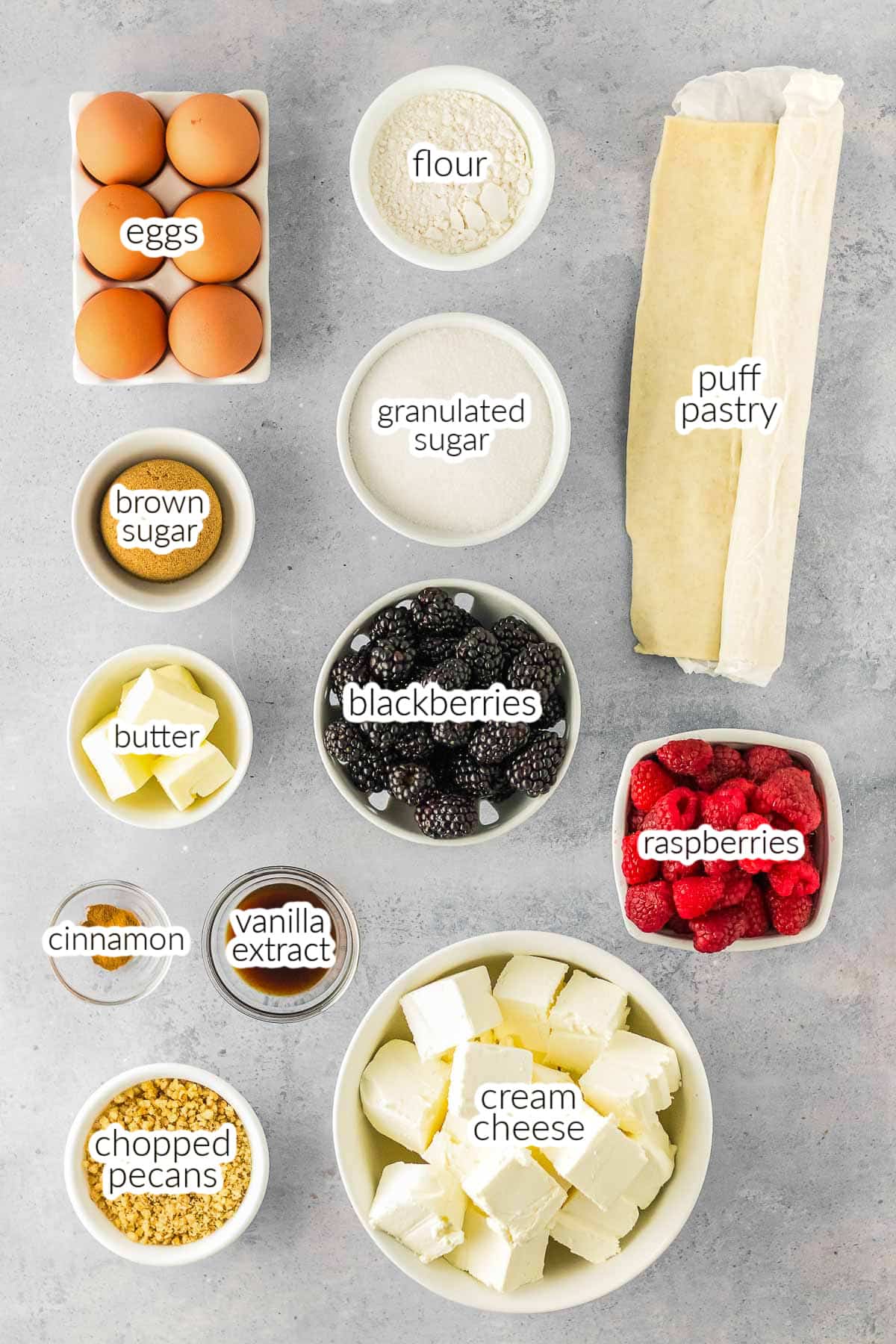 Ingredients for Cream Cheese Puff Pastry arranged on a countertop, including eggs, flour, puff pastry, sugars, butter, blackberries, raspberries, cinnamon, vanilla, pecans, and cream cheese.