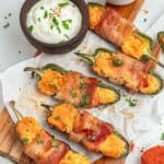 Grilled jalapeño poppers stuffed with cheese and wrapped in bacon are arranged on a wooden board, served with a bowl of sour cream and a small dish of chopped herbs.