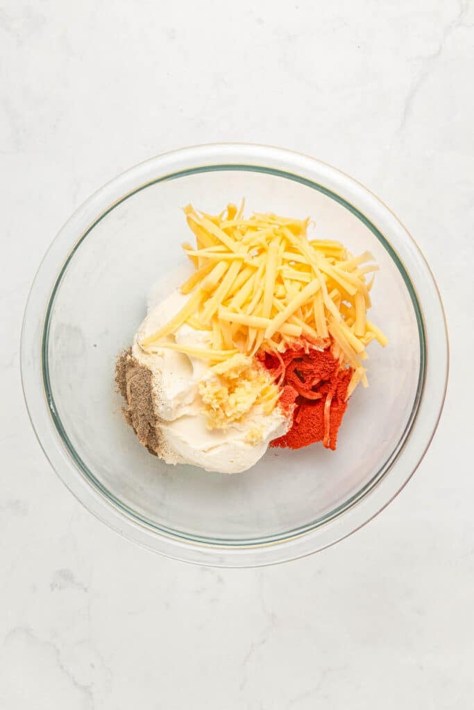 A glass bowl containing shredded cheese, cream cheese, minced garlic, paprika, black pepper, and onion powder on a white surface.