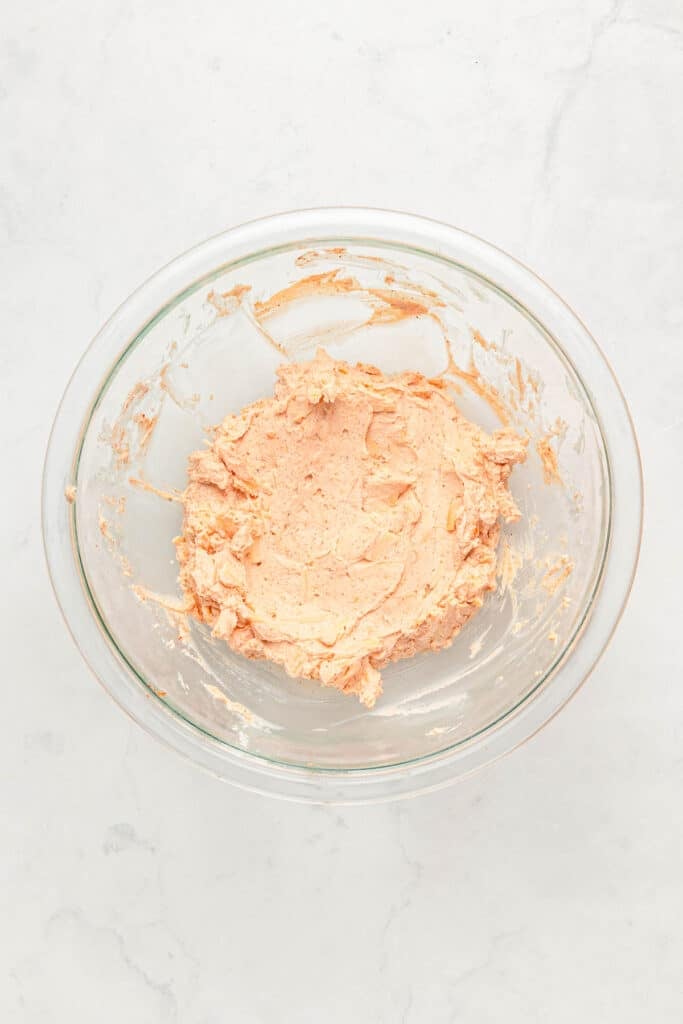A glass mixing bowl containing a creamy jalapeño filling mixture sits on a white marble surface.