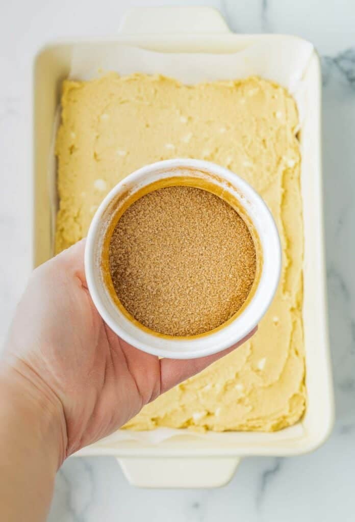 A hand holds a small bowl of brown sugar cinnamon sugar above an unbaked cookie batter in a rectangular baking dish.