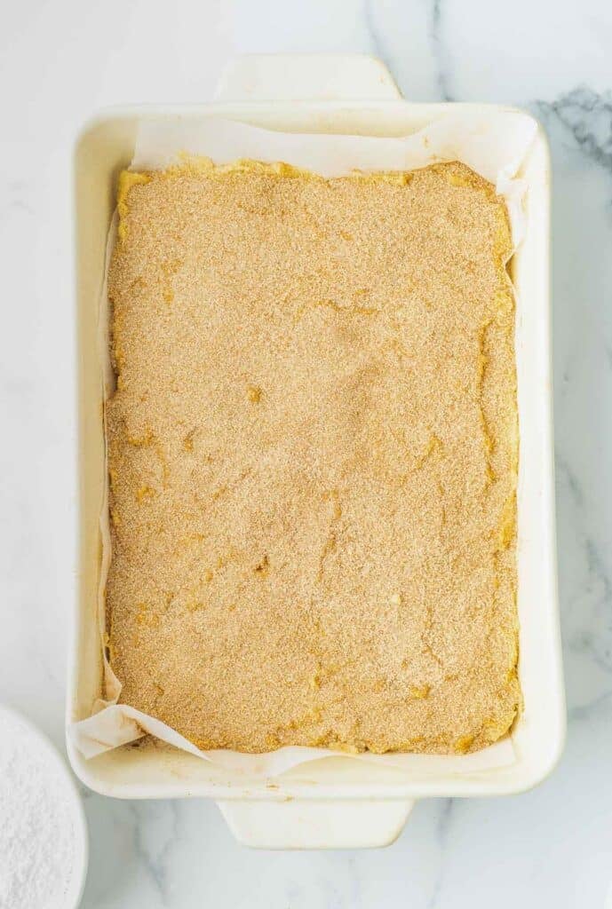 A rectangular baking dish lined with parchment paper, filled with an even layer of unbaked crumbly dough mixture.