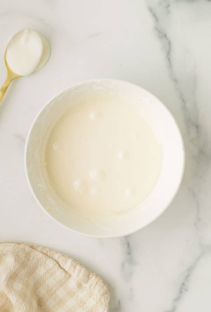 A white bowl filled with smooth white icing sits on a marble surface next to a gold spoon with icing and a beige textured cloth.