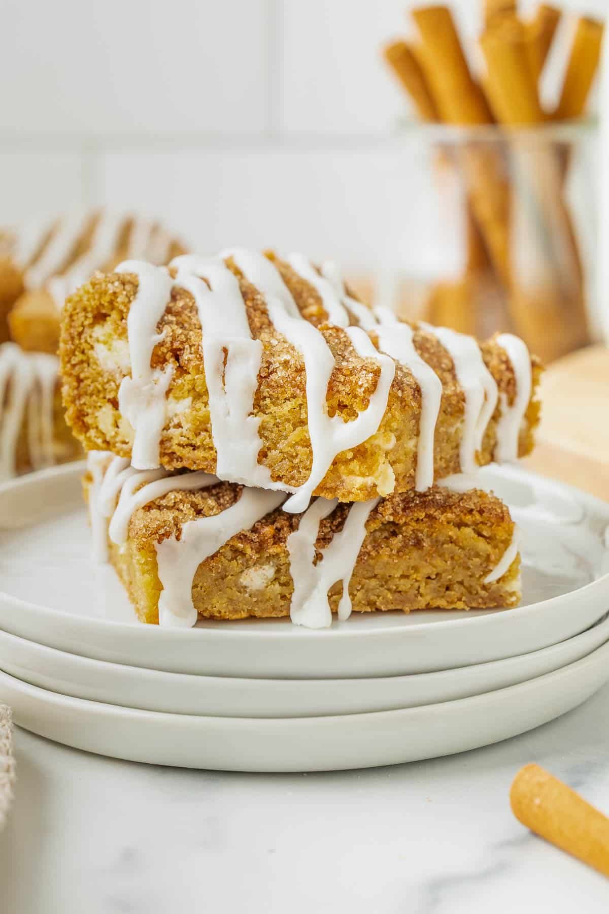 Two pieces of Snickerdoodle Cookie Bars stacked on white plates, topped with white icing drizzle. Cinnamon sticks are visible in the background.
