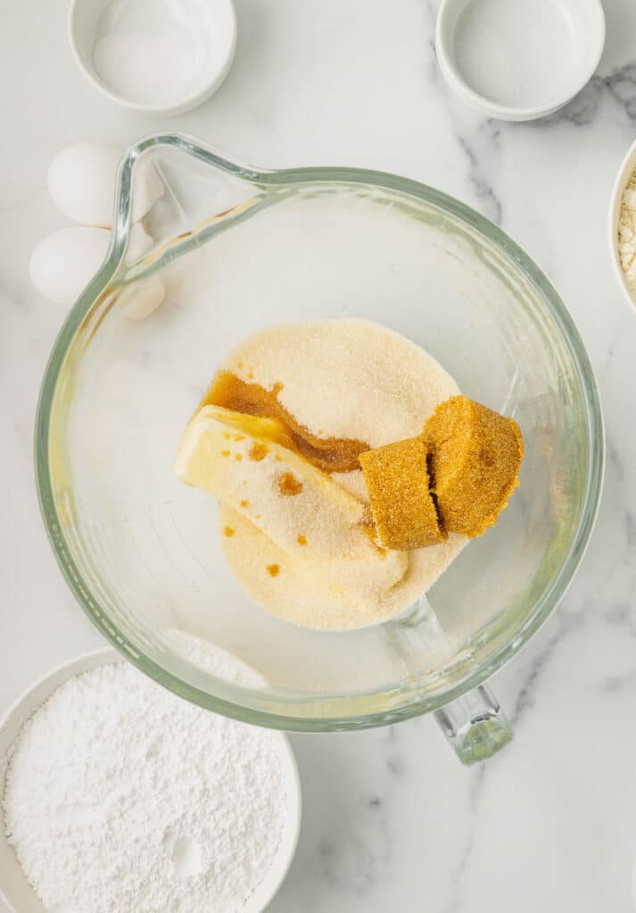 A glass mixing bowl with butter, brown sugar, granulated sugar, and vanilla on a white surface, surrounded by ingredients including powdered sugar and eggs.