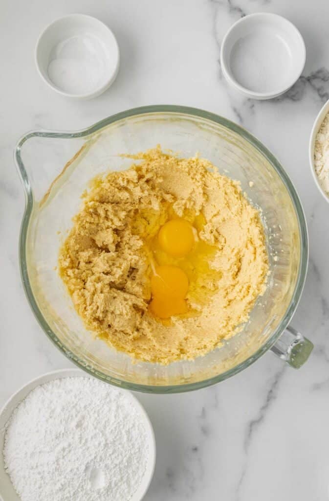 A glass mixing bowl with creamed sugar and butter, two raw eggs on top, surrounded by bowls of flour, powdered sugar, and baking ingredients on a white countertop.