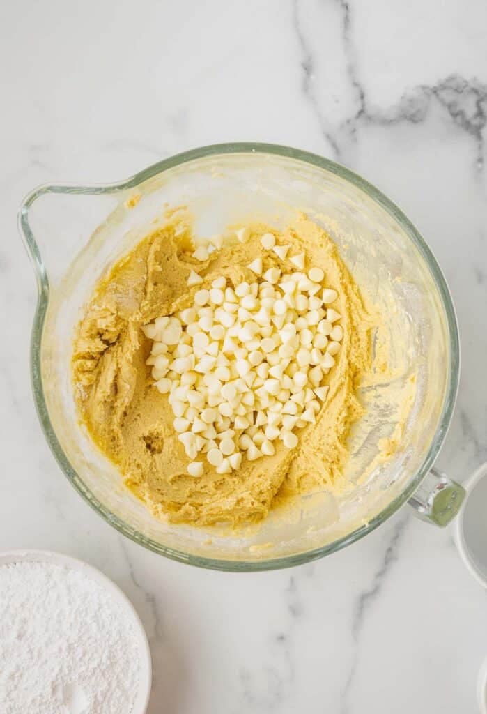 A glass mixing bowl on a marble surface contains cookie dough with white chocolate chips on top, ready to be mixed in.