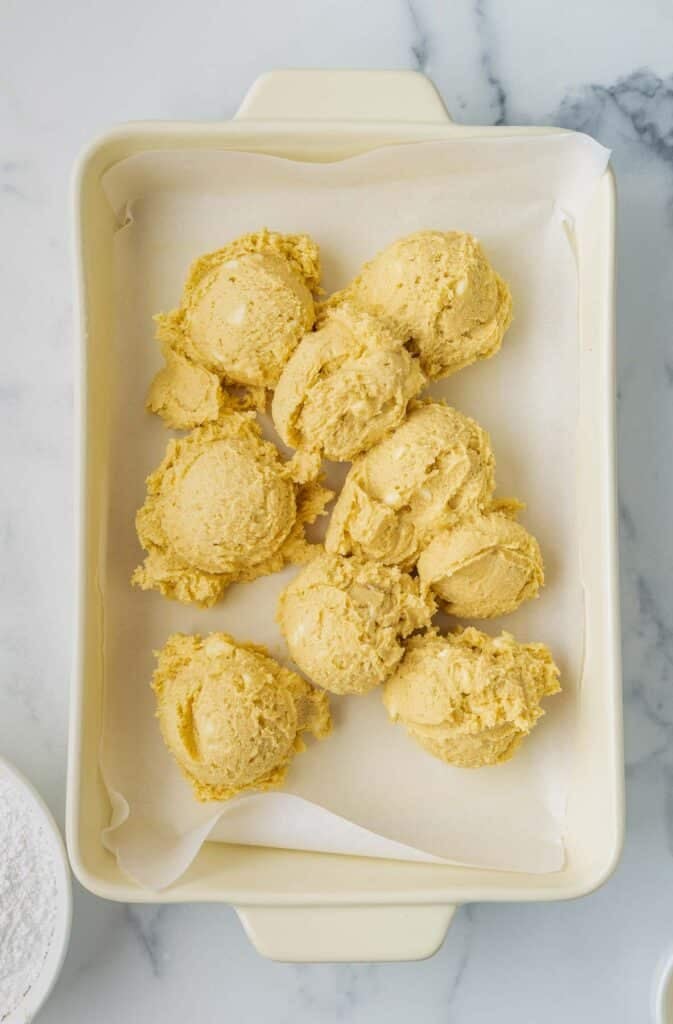 A parchment-lined baking dish with seven scoops of raw cookie dough on a white marble surface.