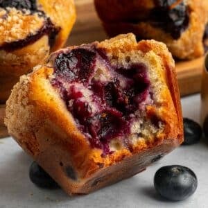 Close-up of a blueberry muffin with a bite taken out, showing the moist interior and visible blueberries, with whole blueberries and other muffins in the background.