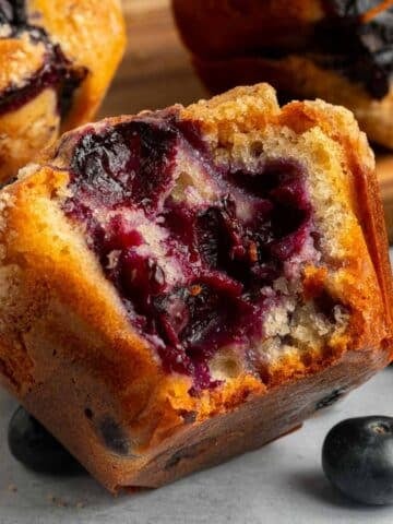 Close-up of a blueberry muffin with a bite taken out, showing the moist interior and visible blueberries, with whole blueberries and other muffins in the background.