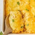 A wooden spoon lifts a serving of cheesy casserole topped with melted cheese and garnished with cilantro from a baking dish.