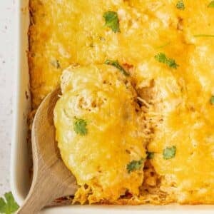 A wooden spoon lifts a serving of cheesy casserole topped with melted cheese and garnished with cilantro from a baking dish.