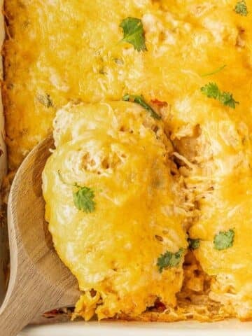 A wooden spoon lifts a serving of cheesy casserole topped with melted cheese and garnished with cilantro from a baking dish.