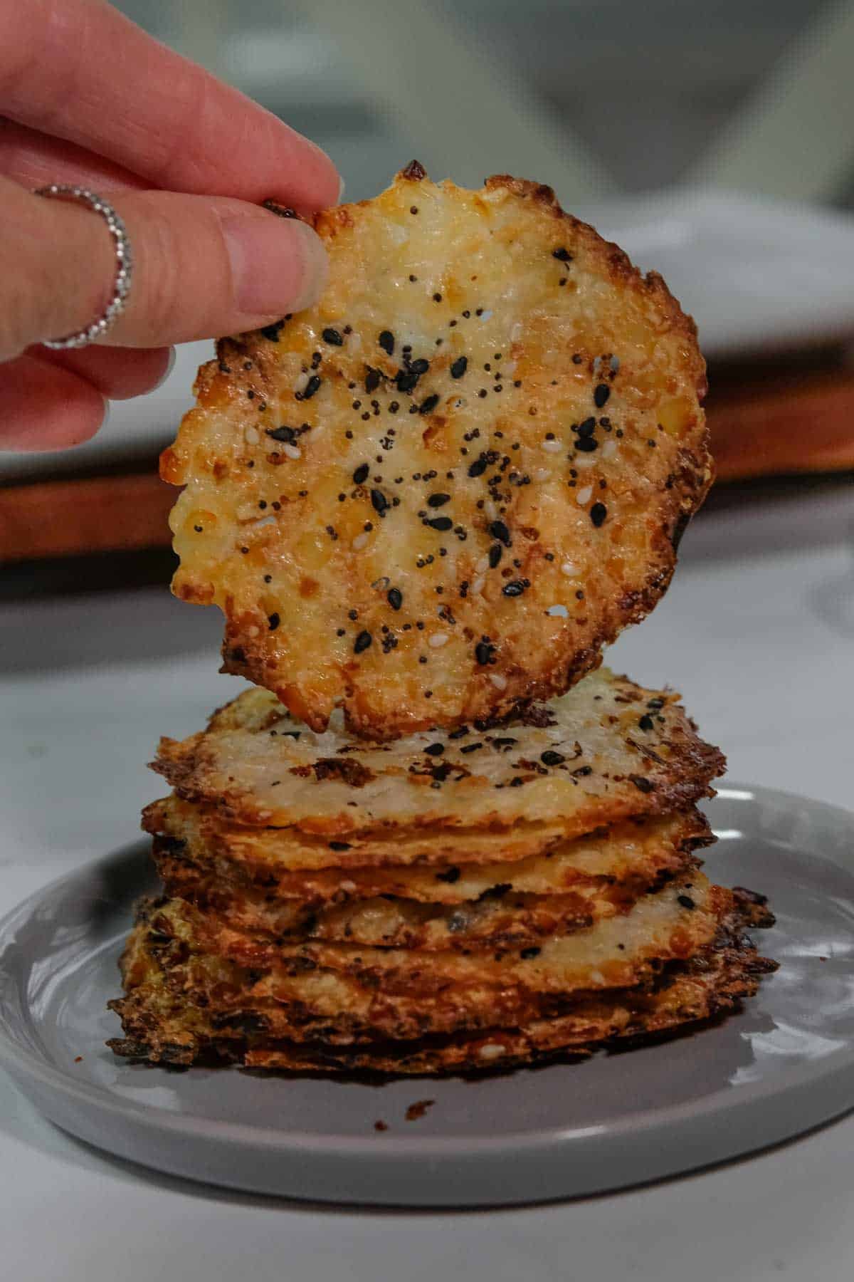A hand holds a round, crispy cheese chip topped with sesame seeds above a stack of similar chips on a plate.