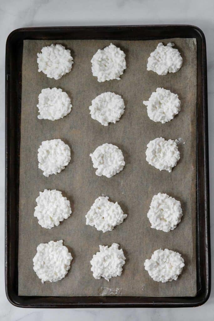 A baking tray lined with parchment paper holds fifteen evenly spaced dollops of a cottage cheese.