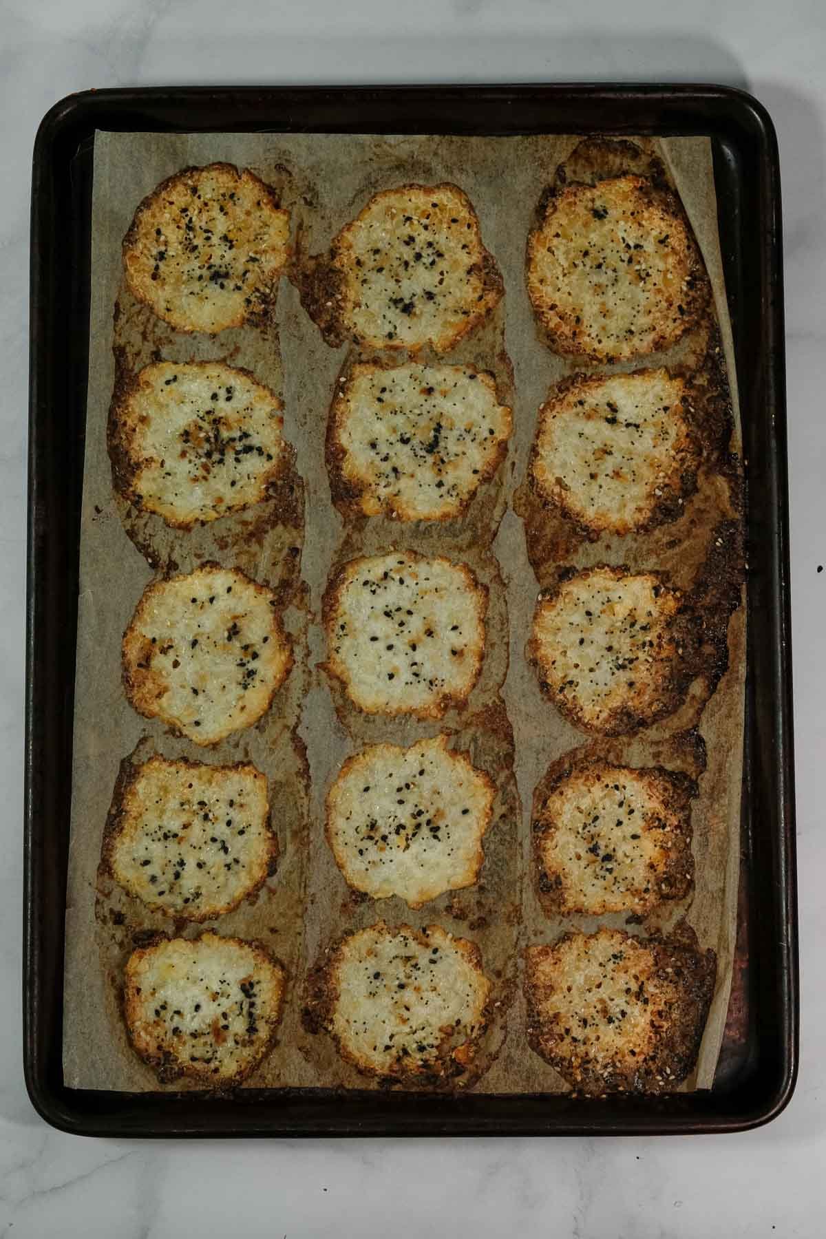 A baking tray lined with parchment paper holds 15 round, golden-brown cottage cheese chips topped with everything bagel seasoning.