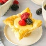 A cream cheese puff pastry filled with cream filling topped with two raspberries and one blackberry, served on a small white plate.