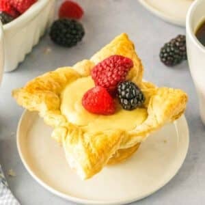 A cream cheese puff pastry filled with cream filling topped with two raspberries and one blackberry, served on a small white plate.
